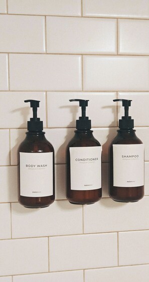 Tre bottiglie di bagnoschiuma, balsamo e shampoo della Collezione Margaux, disposte su un muro piastrellato.