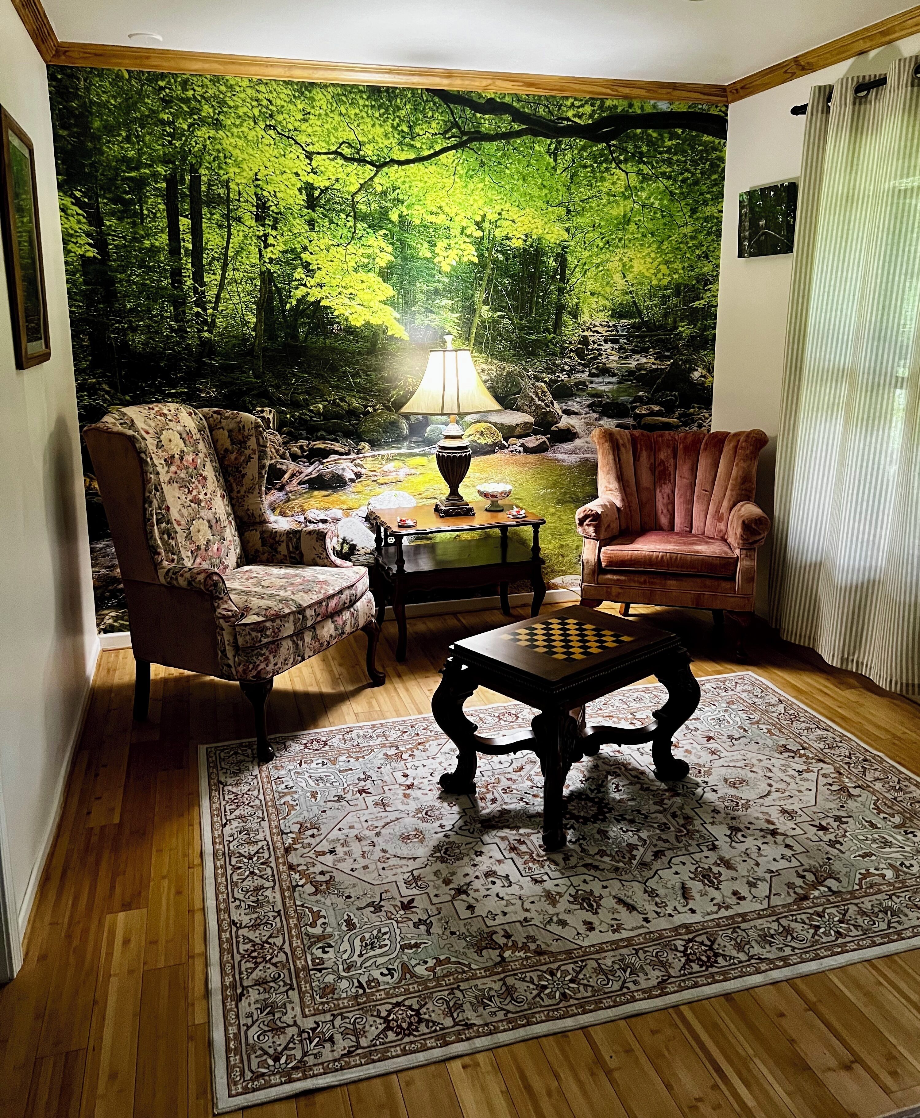 Ein Wohnzimmer mit einer großen Waldmalerei, zwei Sesseln, einem Couchtisch und einem Teppich.
