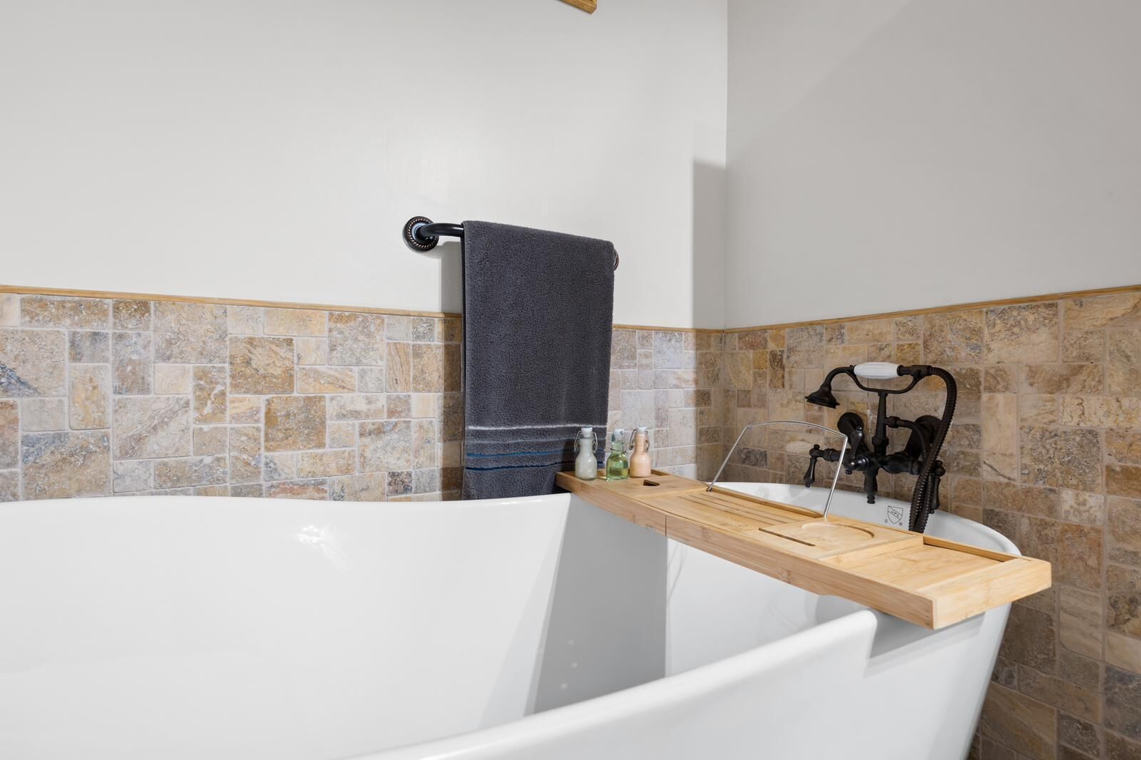 A bathroom with a bathtub, a wooden shelf, a towel rack, and a stone-tiled wall.