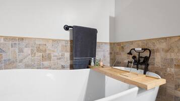 A bathroom with a bathtub, a wooden shelf, a towel rack, and a stone-tiled wall.