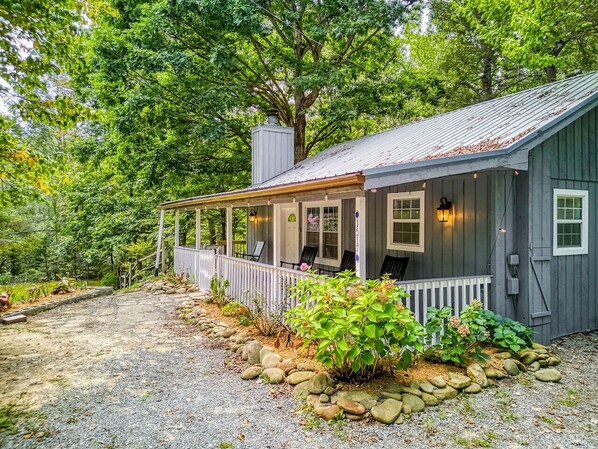 A rustic cabin with a porch, surrounded by a gravel driveway and a stone border.