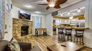 Un salon moderne avec une cheminée en pierre, un ventilateur de plafond et un coin bar avec des tabourets.