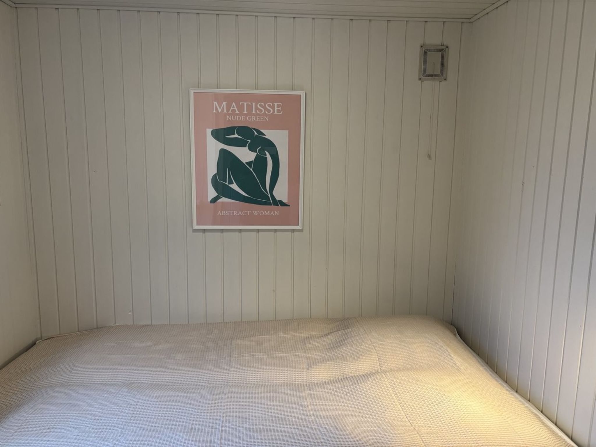 Une pièce avec un lit, une affiche représentant un tableau accrochée au mur et une petite grille d’aération.