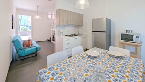 A compact kitchen with a stove, microwave, and refrigerator. There is a dining table with chairs and a patterned tablecloth. A blue chair is visible in the background.