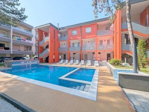 Une piscine avec des chaises longues, un immeuble à plusieurs étages doté de balcons et un ciel dégagé.