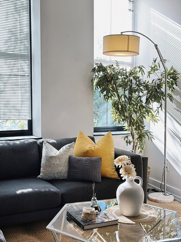 A modern living room with a dark sofa, yellow cushions, a glass coffee table, a floor lamp, and a large plant.