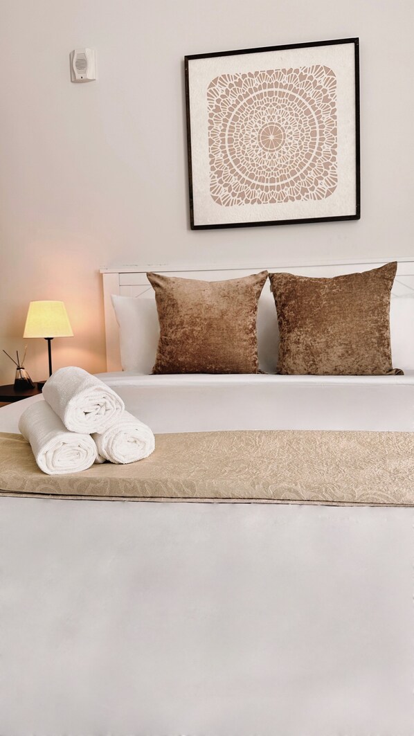 Een net opgemaakt bed met witte lakens en twee bruine kussens, een lijst met abstract kunstwerk erboven en een opgerolde witte handdoek aan de rand van het bed.