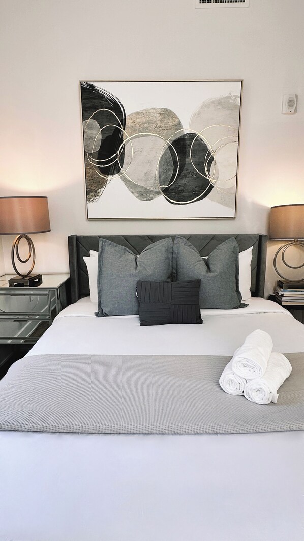 A hotel room with a large bed, grey bedding, a grey headboard, and a modern abstract painting above the bed.