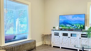 A living room with a large window, a flat-screen TV, a white cabinet, and a glass table. - Light filled apt in Historic Home by UW & Downtown (Seattle)