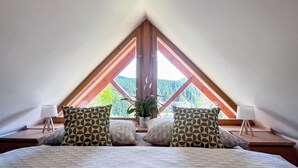 A bedroom with a sloped ceiling, a bed with patterned bedding, two pillows, a bedside lamp, and a view of a forest through a window. - Sachrův Kopec by Interhome (Harrachov)