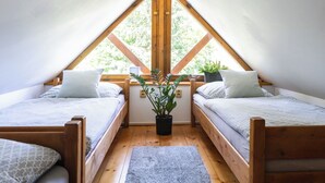 Une petite chambre mansardée avec deux lits en bois, une plante en pot et une vue sur les arbres à travers une grande fenêtre triangulaire.
