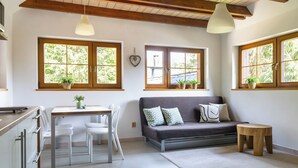 Une cuisine moderne avec des meubles blancs, une table à manger avec des chaises, un canapé gris et un petit tabouret en bois. Il y a des fenêtres avec des plantes sur les rebords et une décoration en forme de cœur accrochée au mur.