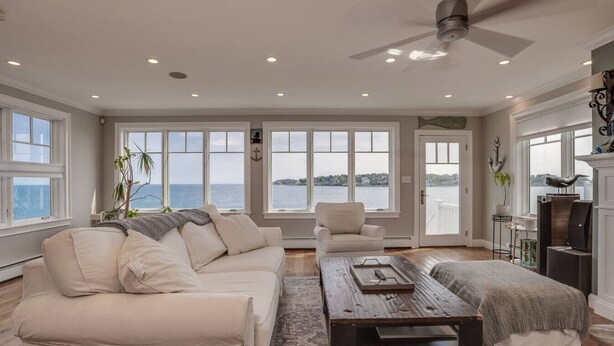 Un salon spacieux avec un grand canapé, une table basse et un fauteuil. De nombreuses fenêtres offrent une vue sur la mer.
