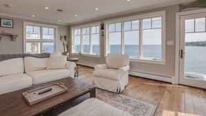 Un salon spacieux avec une grande fenêtre donnant sur la mer, un canapé blanc, un fauteuil blanc et une table basse en bois.