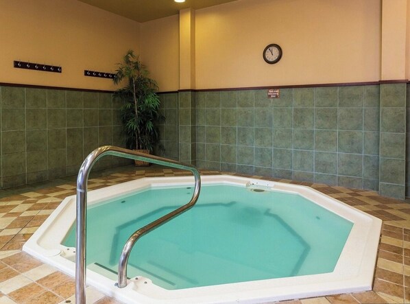 Une petite piscine intérieure avec un garde-corps métallique, entourée de murs recouverts de carreaux verts et d’une horloge fixée au mur.