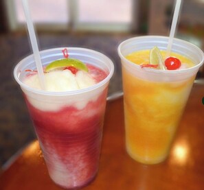 Dos vasos de plástico con pajitas, uno con una bebida roja y otro con una bebida naranja, ambos decorados con frutas.