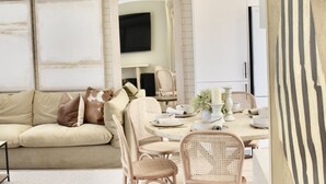 Una sala de estar moderna con un sofá beige, sillas de mimbre, una mesa de comedor preparada para cuatro comensales y un televisor de pantalla plana montado en la pared.