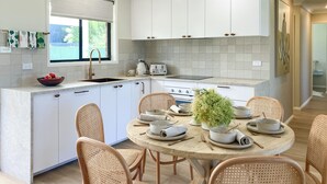 Una cocina moderna con armarios blancos, una mesa redonda de comedor, sillas de mimbre y un ventanal con vista a follaje.