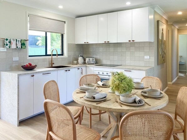 A modern kitchen with white cabinets, a round dining table, wicker chairs, and a window with a view of greenery.