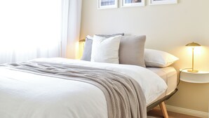 Una cama bien hecha con una manta blanca y beige, una lámpara de noche y tres cuadros en la pared.