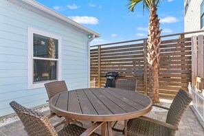 A patio with a round wooden table and wicker chairs, a grill, and a palm tree.