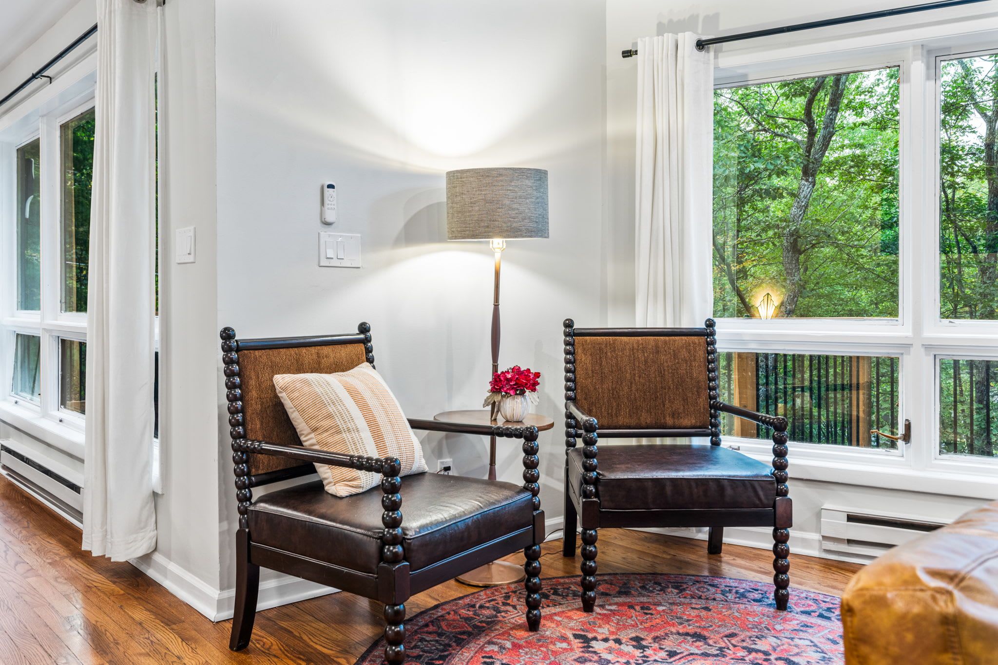 A room with two wooden chairs, a lamp, and a view of trees through large windows.