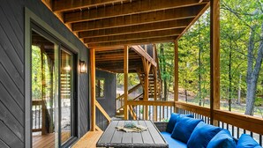 A wooden deck with a sofa and a table, surrounded by glass doors and windows, overlooking a forested area.