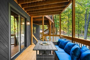 A wooden deck with a sofa and a table, surrounded by glass doors and windows, overlooking a forested area. - Pocono Speak Easy Hot Tub Golf Simulator 1920 Bar (East Stroudsburg)