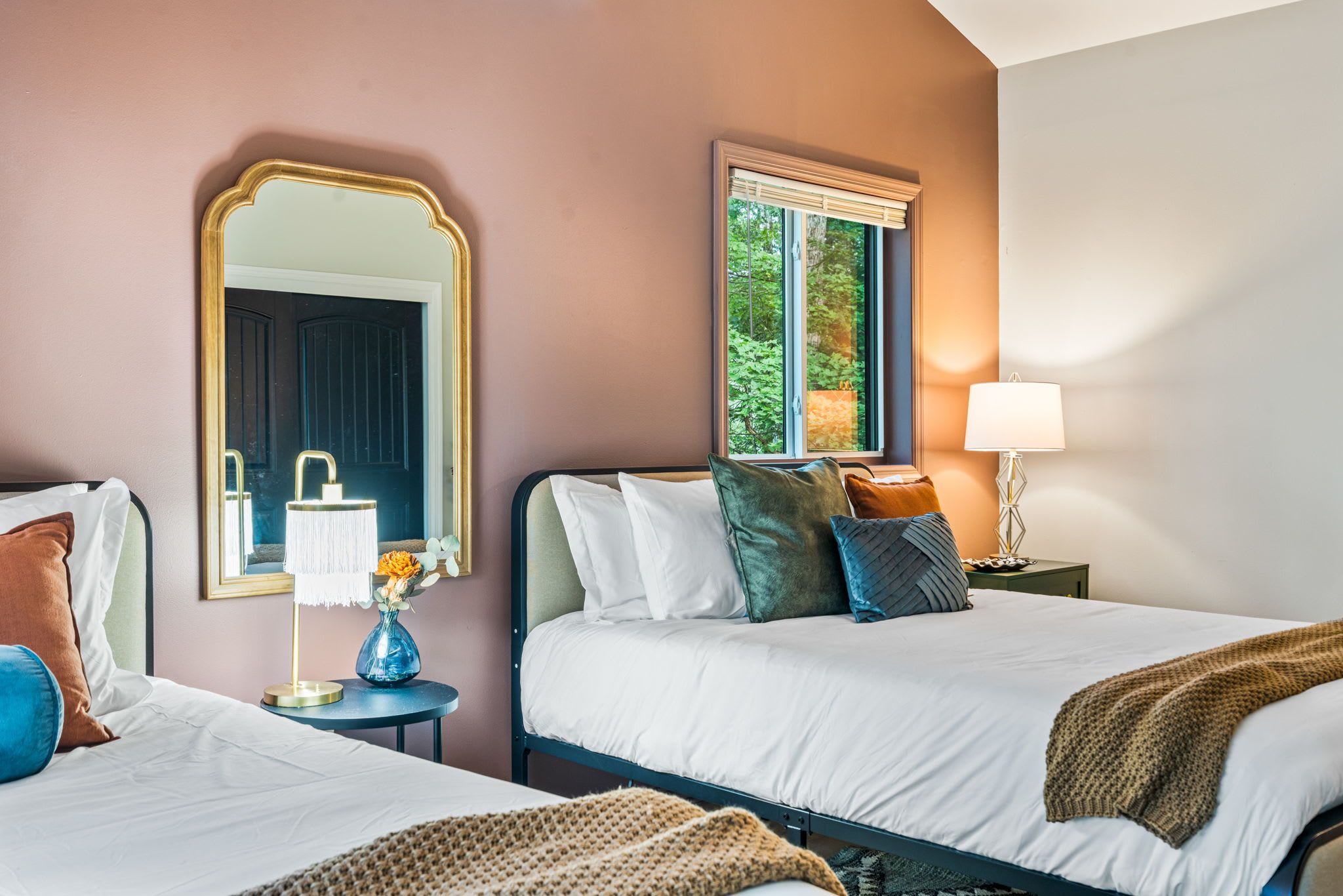 A hotel room with two beds, a large mirror, a window with a view of greenery, and a bedside table with a lamp and vase.