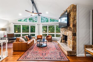 A spacious living room with a stone fireplace, large windows, and a cozy seating area. - Pocono Speak Easy Hot Tub Golf Simulator 1920 Bar (East Stroudsburg)