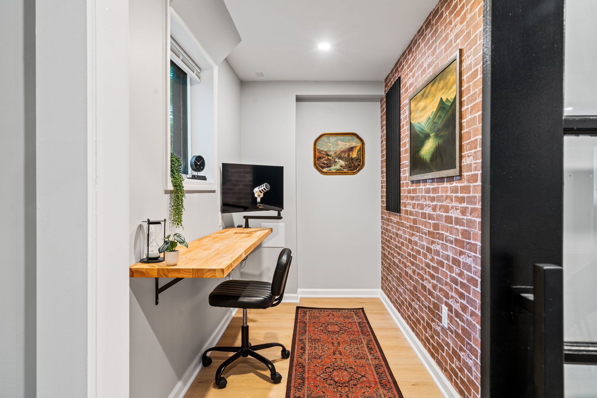 Un couloir étroit avec un bureau en bois, une chaise de bureau noire et un écran d’ordinateur. Un tapis rouge à motifs est posé sur le sol, et deux tableaux encadrés ornent le mur de briques.