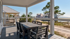 Un espace extérieur aménagé, abrité et doté de tables et de chaises, offrant une vue sur une plage bordée de maisons et d’arbres.