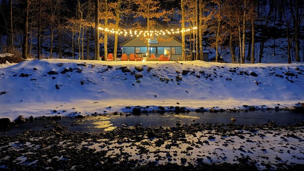 Una cabaña en un paisaje nevado, rodeada de árboles, con luces colgantes sobre la entrada.
