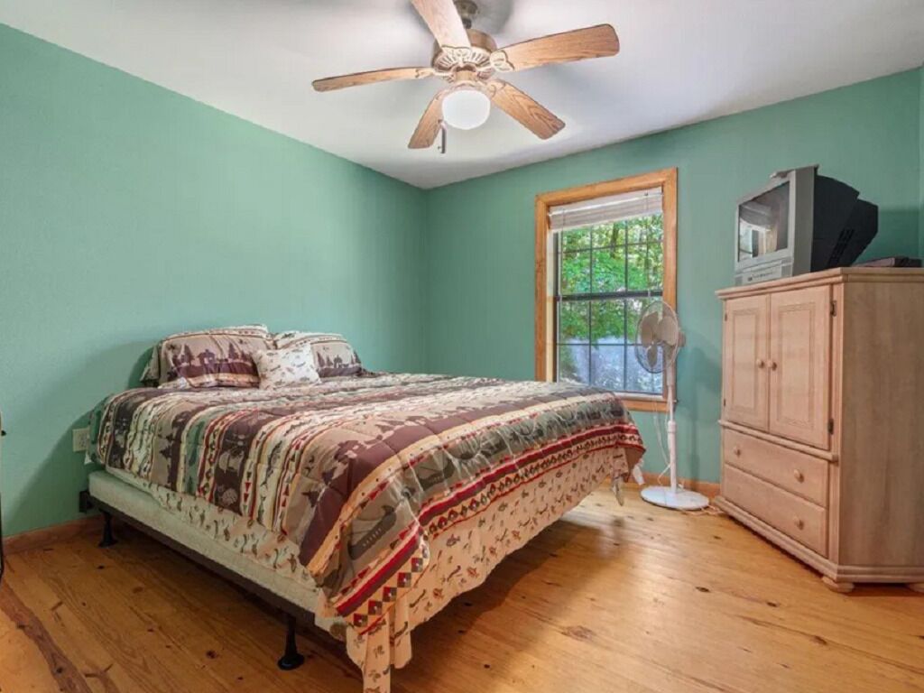 Un dormitorio con cama, suelo de madera, ventilador de techo, ventana y armario de madera.
