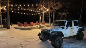 Un vehículo todoterreno blanco estacionado en un espacio exterior nevado con un hogar de piedra y sillas de madera bajo luces colgantes.