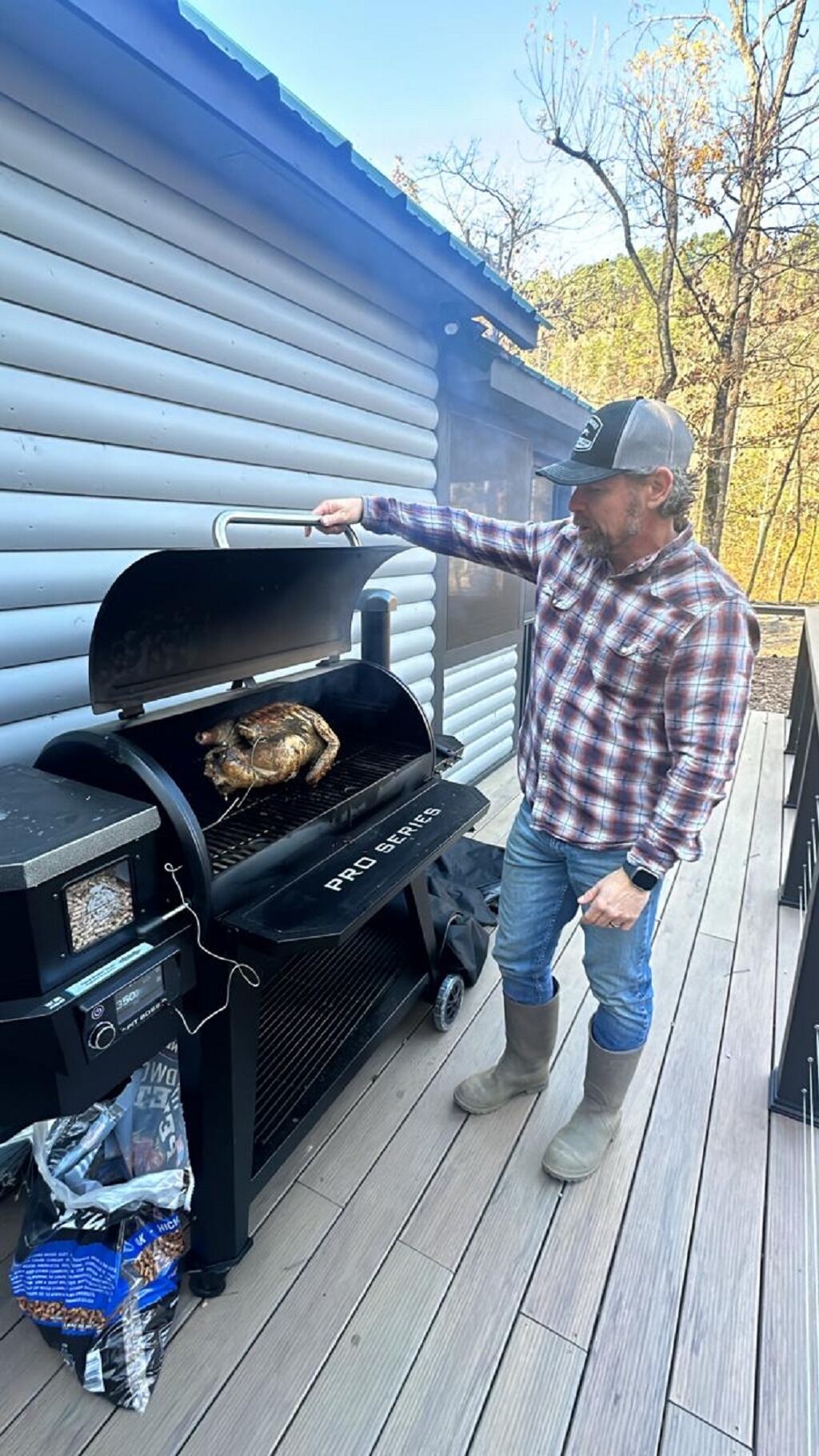 A man grilling outdoors on a deck with a portable grill, a bag of charcoal, and a blue building in the background.