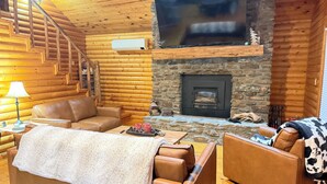 Un acogedor interior de cabaña de madera con chimenea, sofás de cuero y un televisor colocado sobre la chimenea.
