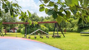 A playground with a swing set, a slide, and a trampoline in a grassy area with trees. - 6 Person Holiday Park Home in Grindsted (Grindsted)