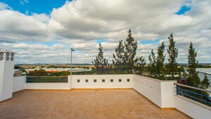 A rooftop terrace with a tiled floor, a railing, and a view of trees and buildings in the distance. - Urbaniza o da Pegada Apartamento T2 (Tavira)