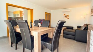 A dining area with a wooden table and black chairs, a sofa, and a kitchen in the background. - Urbaniza o da Pegada Apartamento T2 (Tavira)