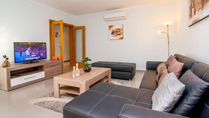A modern living room with a television, a wooden coffee table, a black leather sofa, and a small potted plant. - Urbaniza o da Pegada Apartamento T2 (Tavira)