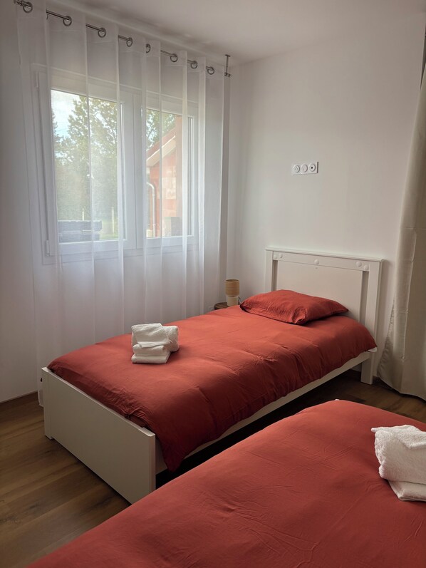 Ein Schlafzimmer mit zwei Einzelbetten, weißen Vorhängen, einem Nachttisch mit Lampe und einem Fenster mit Blick auf Bäume und ein Gebäude.