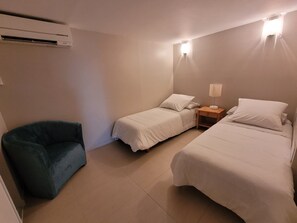Quarto de hotel com duas camas de solteiro, uma cadeira, uma mesa de cabeceira com abajur e um aparelho de ar condicionado.