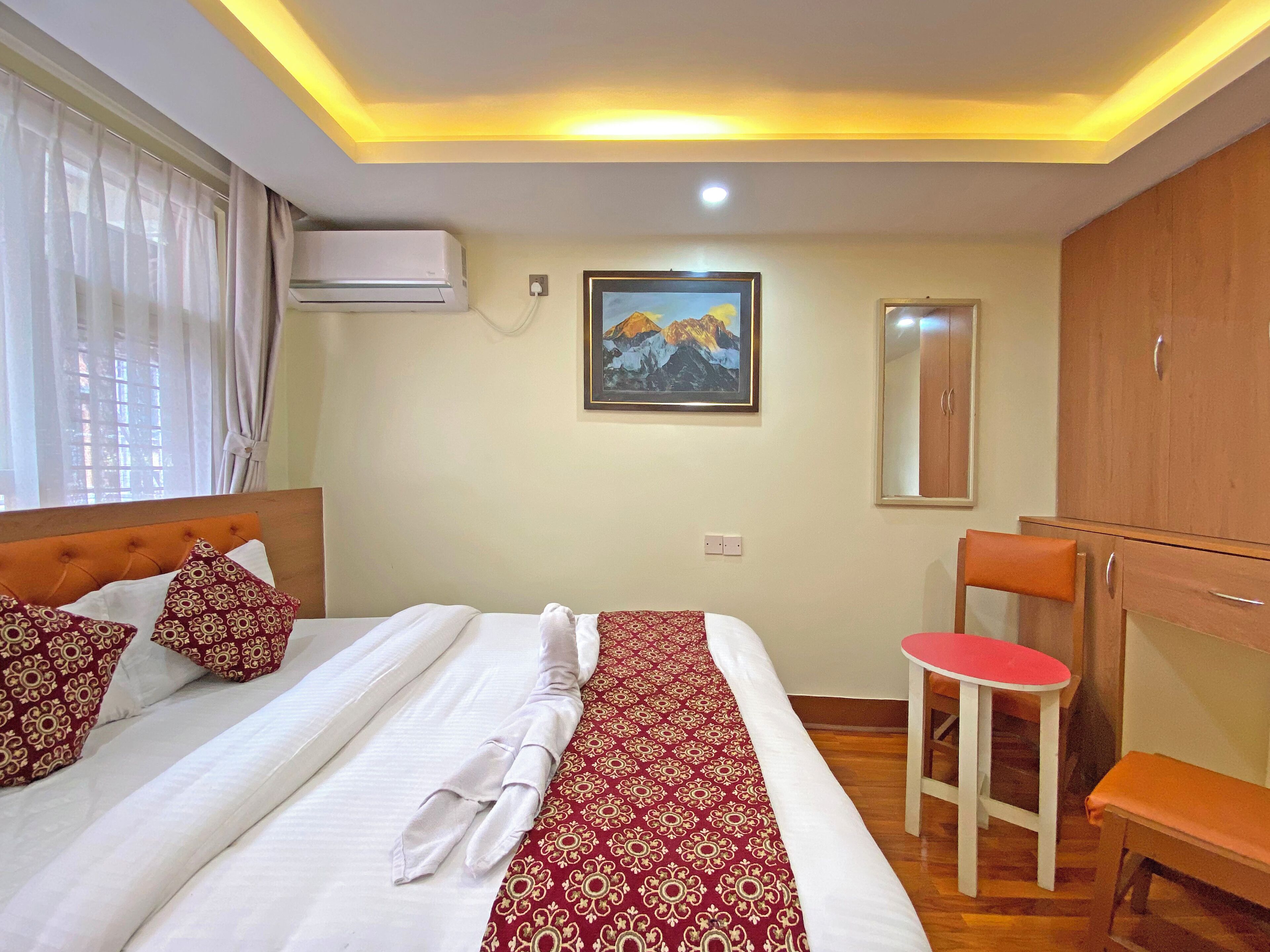 A hotel room with a neatly made bed, a red and white patterned bedspread, a painting of mountains on the wall, a wooden wardrobe, and a small stool with a red seat.