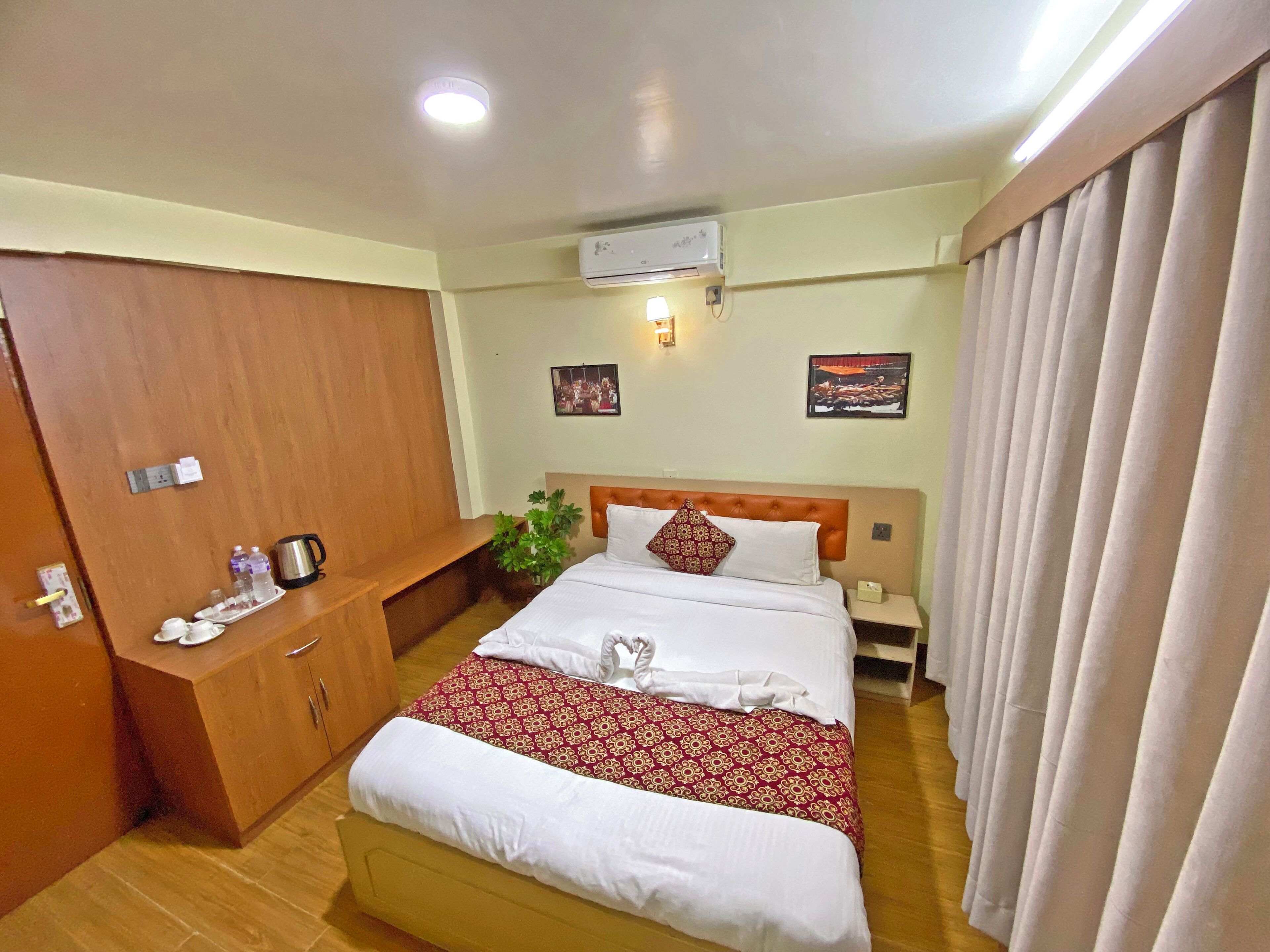 A hotel room with a bed, a wooden wardrobe, a bedside table with a lamp, and a wall-mounted air conditioner.