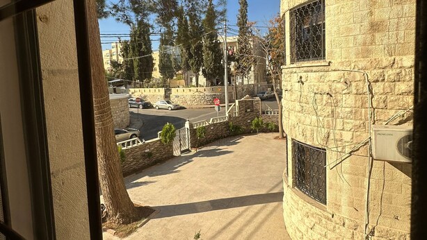 A view through a window of a building with a stone facade, a small tree, and a parking area with cars.