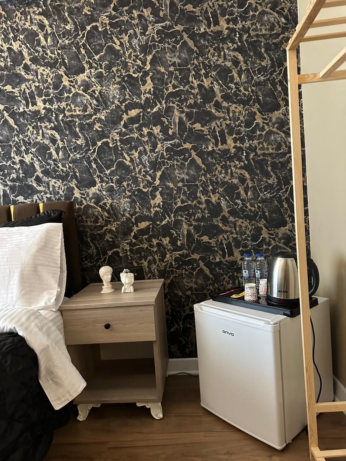 Camera da letto con parete effetto marmo, comodino in legno con due oggetti decorativi, piccolo frigorifero bianco e letto con coperta bianca.