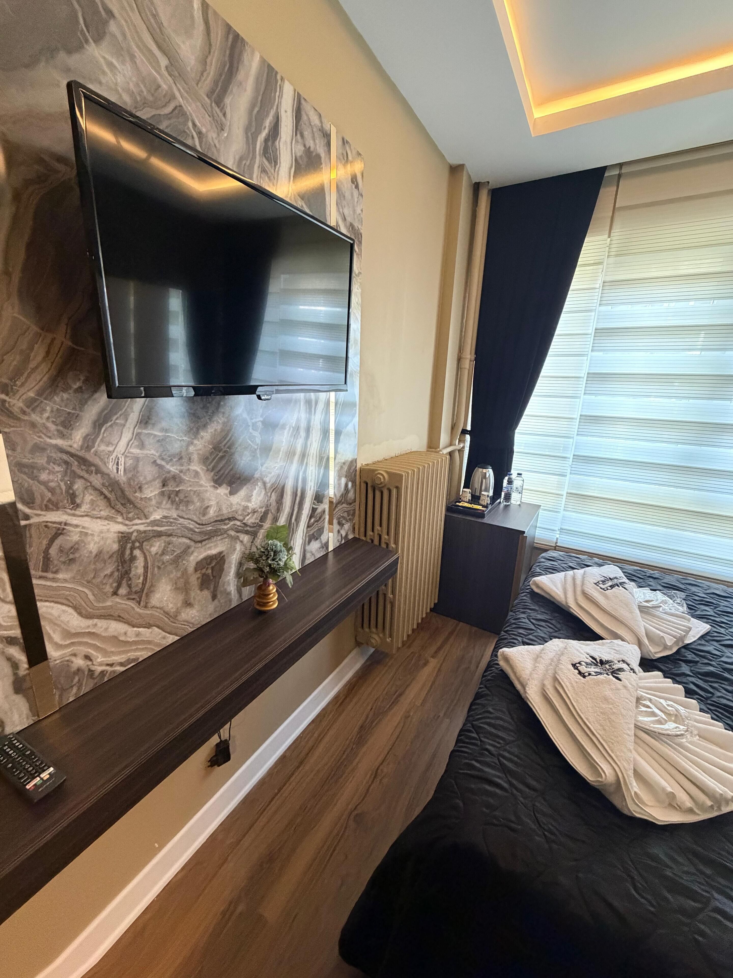 A modern hotel room with a marble wall, a flat-screen TV, a wooden console table, a radiator, and a bed with towels.