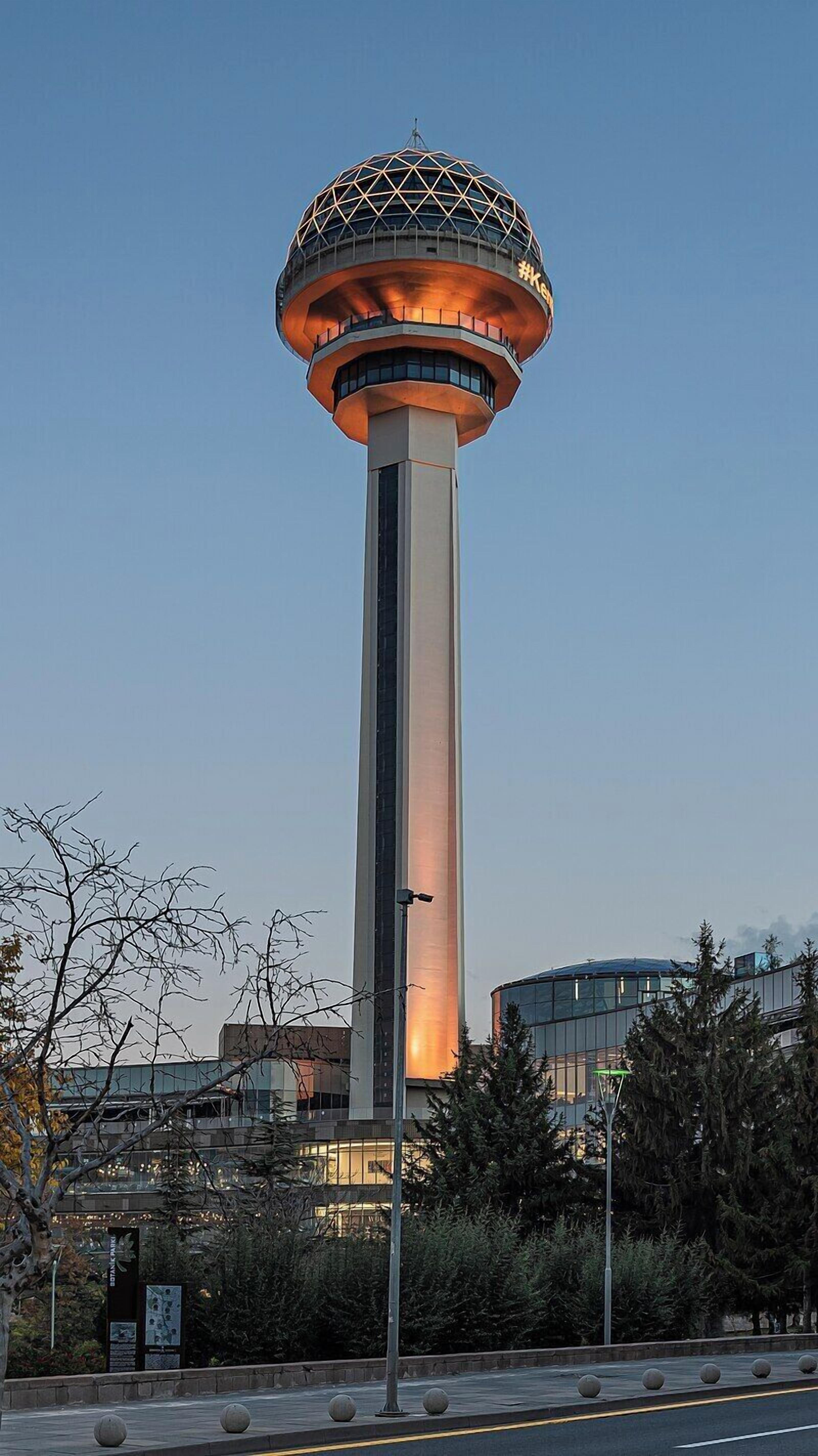 Una torre alta con una plataforma de observación esférica, rodeada de árboles y una calle con bolardos.