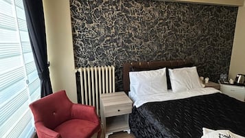 A hotel room with a bed, a red armchair, a small table with cups, a radiator, and a headboard with a textured design.